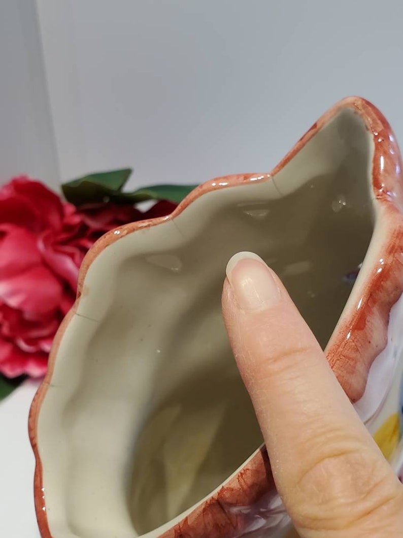May include: A white ceramic planter shaped like a flower with pink, red, and yellow painted details. The planter has a scalloped rim and a smooth, white interior.