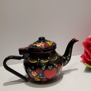 Könnte beinhalten: Schwarze Teekanne mit rotem Herzen und Blumenmuster. Die Teekanne hat einen Deckel und einen Griff. Das Blumenmuster ist in Rot-, Gelb-, Blau- und Grüntönen gehalten.