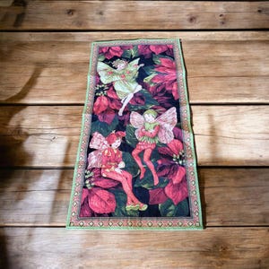 May include: A tapestry wall hanging with a black background and a green border. The tapestry features three fairies with wings, wearing red and green clothing, flying over red flowers.