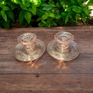 May include: Two pink glass candle holders with etched floral designs. The holders are on a wooden surface.