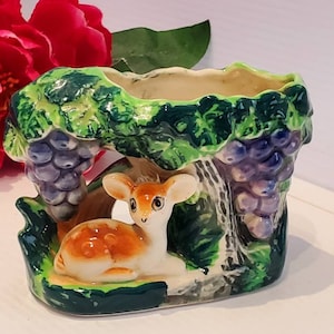 Puede incluir: Un macetero de cerámica con forma de vid con un cervatillo sentado debajo. El macetero está pintado en tonos de verde, marrón y morado.