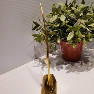 May include: A gold metal sculpture of a sailboat with a long, thin mast and a rounded base. The sculpture is on a white surface.