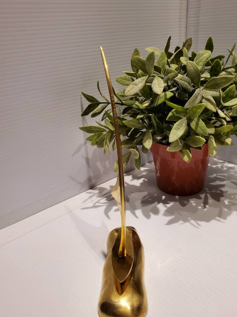 May include: A gold metal sculpture of a sailboat with a long, thin mast and a rounded base. The sculpture is on a white surface.