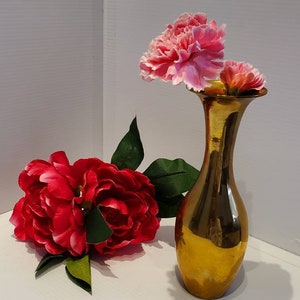 May include: A gold metal vase with a flared opening holds pink and red carnations. The vase is sitting on a white surface.