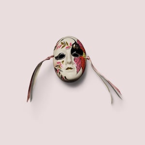 Peut inclure: Un masque en céramique blanche avec des motifs floraux roses, rouges et dorés. Le masque a un œil noir et un motif de papillon noir. Le masque est attaché à deux rubans roses et blancs.