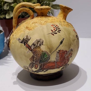 May include: A ceramic pitcher with a round body and two spouts. The pitcher is decorated with a hand-painted scene of a warrior riding a chariot pulled by two horses. The scene is painted in shades of red, green, black, and white.