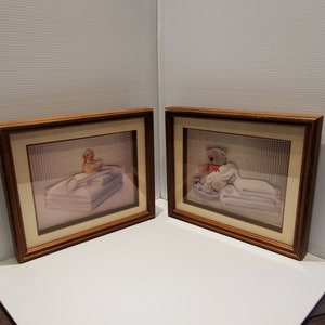 May include: Two framed prints with wooden frames. One print features a yellow duck on a stack of white towels. The other print shows a koala bear with a red bow on a stack of white towels. Both prints have a beige mount.