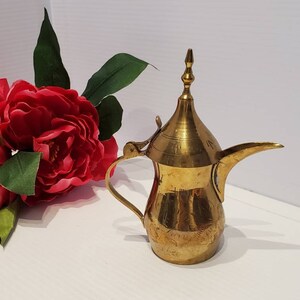 Könnte beinhalten: Eine kleine, goldfarbene Messingkanne mit einem dekorativen Griff und einem spitzen Deckel. Die Kanne hat ein aufwendig graviertes Design.