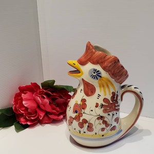 May include: A ceramic pitcher shaped like a rooster with a red comb, yellow beak, and blue eyes. The pitcher is decorated with red, yellow, and blue floral designs.