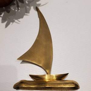 May include: A gold-colored metal sailboat sculpture. The sailboat has a single sail and a small hull. The sculpture is on a white surface.