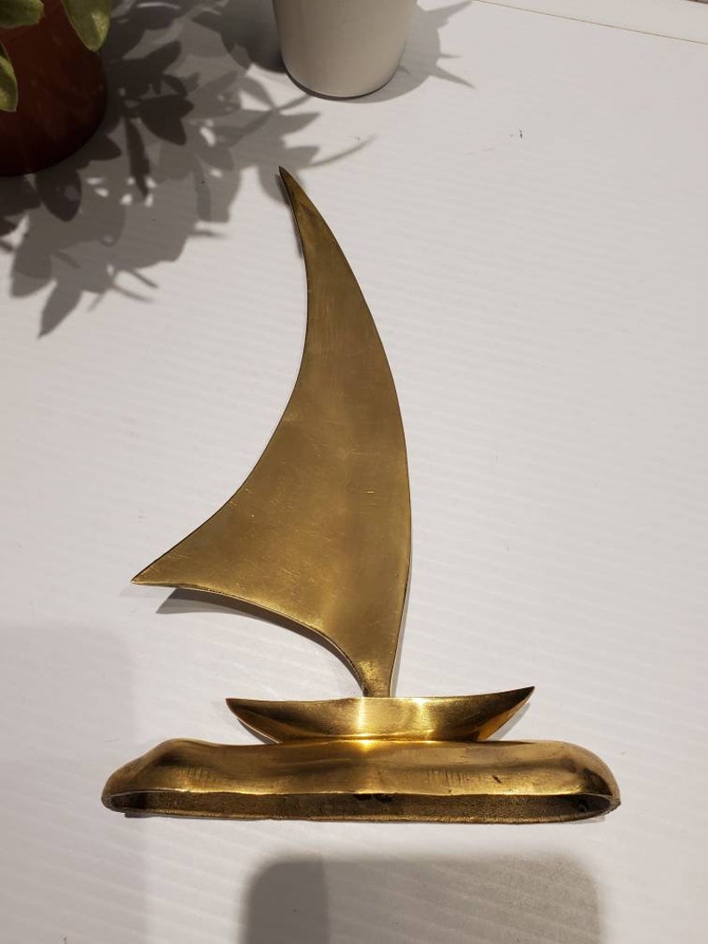 May include: A gold-colored metal sailboat sculpture. The sailboat has a single sail and a small hull. The sculpture is on a white surface.