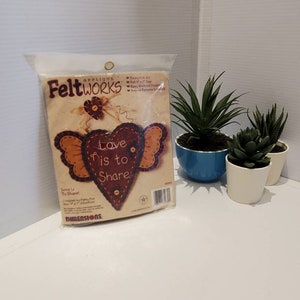 May include: A package of Feltworks applique kit with a heart design that reads "Love is to Share". The kit includes felt pieces and instructions for an easy weekend project. The package is next to three small potted succulents.