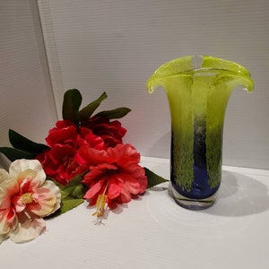 May include: A green and blue glass vase with a unique, wavy design. The vase is sitting on a white surface with red and white flowers.
