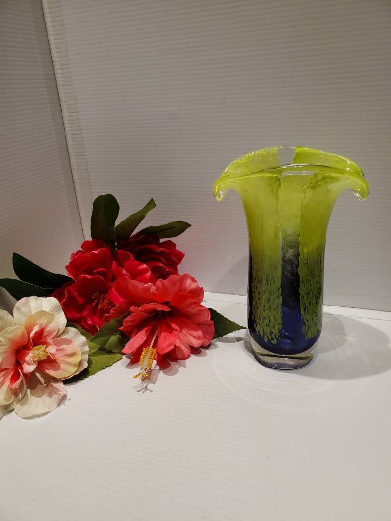May include: A green and blue glass vase with a unique, wavy design. The vase is sitting on a white surface with red and white flowers.