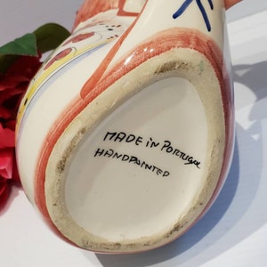 May include: A white ceramic pitcher with a red, blue, and yellow hand-painted design. The bottom of the pitcher reads "MADE in Portugal HANDPAINTED".