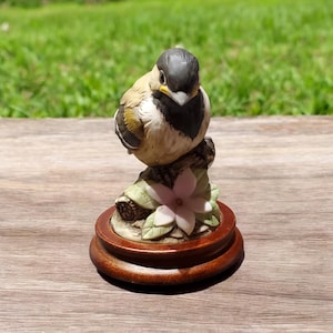 Puede incluir: Una figurita de porcelana de un pájaro pequeño posado en una rama con una flor rosa. El pájaro es marrón, negro y blanco con la cabeza negra. La figurita está sobre una base de madera.