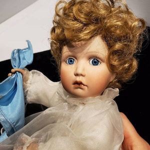 May include: A porcelain doll with blue eyes and curly blonde hair. The doll is wearing a white dress with a sheer overlay and holding a blue cloth. The doll's face has a delicate, painted expression.