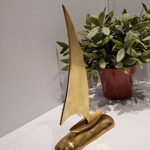 May include: A gold-colored metal sculpture of a sailboat on a base. The sailboat has a tall, pointed sail and a small hull. The base is textured and resembles a rock.