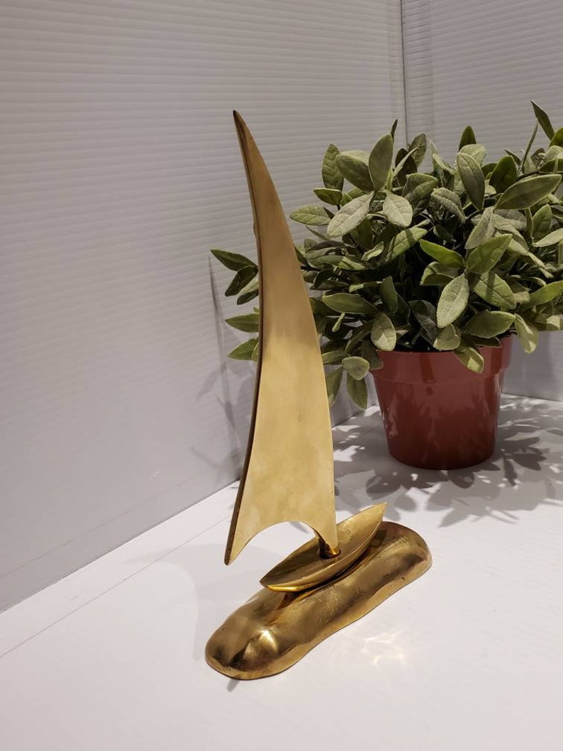 May include: A gold-colored metal sculpture of a sailboat on a base. The sailboat has a tall, pointed sail and a small hull. The base is textured and resembles a rock.