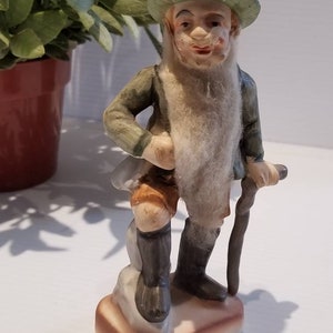 May include: A porcelain figurine of a man with a white beard and a green hat. He is wearing a green jacket, brown shorts, and black boots. He is holding a walking stick and standing on a small base.