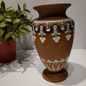 May include: A brown ceramic vase with a wide rim and a pedestal base. The vase is decorated with light blue, white, and brown floral and geometric patterns. The vase is a decorative home decor item.