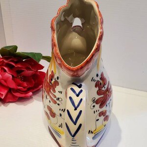 May include: A white ceramic pitcher with a rooster design. The pitcher has red, blue, and yellow accents and a floral pattern. The pitcher has a handle and a spout.