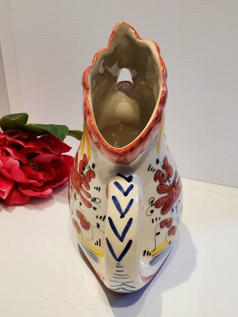 May include: A white ceramic pitcher with a rooster design. The pitcher has red, blue, and yellow accents and a floral pattern. The pitcher has a handle and a spout.