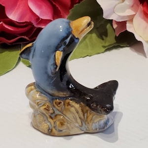 May include: A ceramic figurine of two dolphins, one blue and one black, leaping out of the water. The dolphins are perched on a brown and tan ceramic base that resembles a wave.