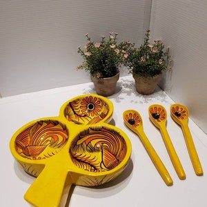 May include: A yellow ceramic serving dish shaped like a clover with three individual bowls. The dish is decorated with orange and black floral designs. Three yellow wooden spoons with black floral designs are included.