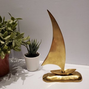 May include: A gold-colored metal sculpture of a sailboat with a single sail. The sailboat is sitting on a base that resembles a wave.
