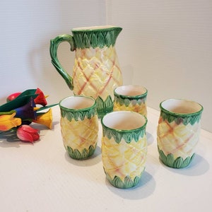 May include: A set of four yellow and green pineapple-themed ceramic glasses and a matching pitcher. The glasses and pitcher have a textured surface that resembles a pineapple.