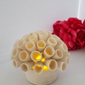 Pode incluir: Uma lâmpada de cerâmica branca em forma de recife de coral com uma luz amarela quente brilhando de dentro. A lâmpada está sobre uma superfície branca.