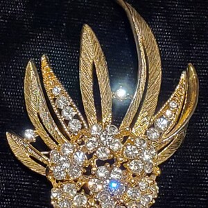 May include: A gold-toned brooch with a cluster of clear rhinestones in the center, surrounded by gold leaves with a textured design.
