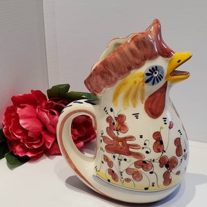 May include: A ceramic pitcher shaped like a rooster with a white base and colorful floral designs in red, yellow, and blue. The rooster's head is raised and its beak is open.