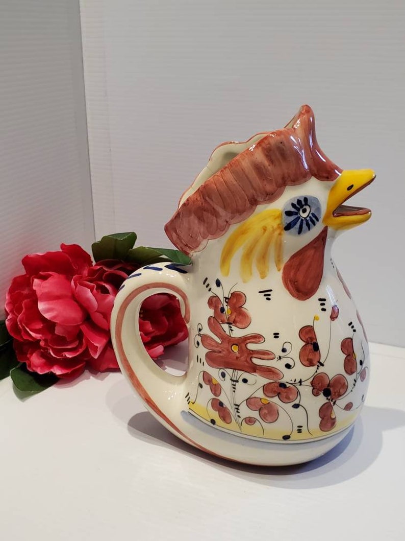 May include: A ceramic pitcher shaped like a rooster with a white base and colorful floral designs in red, yellow, and blue. The rooster's head is raised and its beak is open.