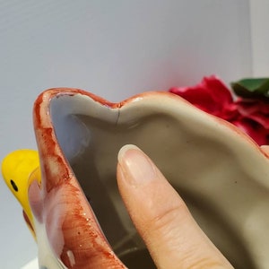 May include: Close-up of the inside of a ceramic rooster planter. The planter is painted in shades of red, yellow, and blue. The inside of the planter is white.