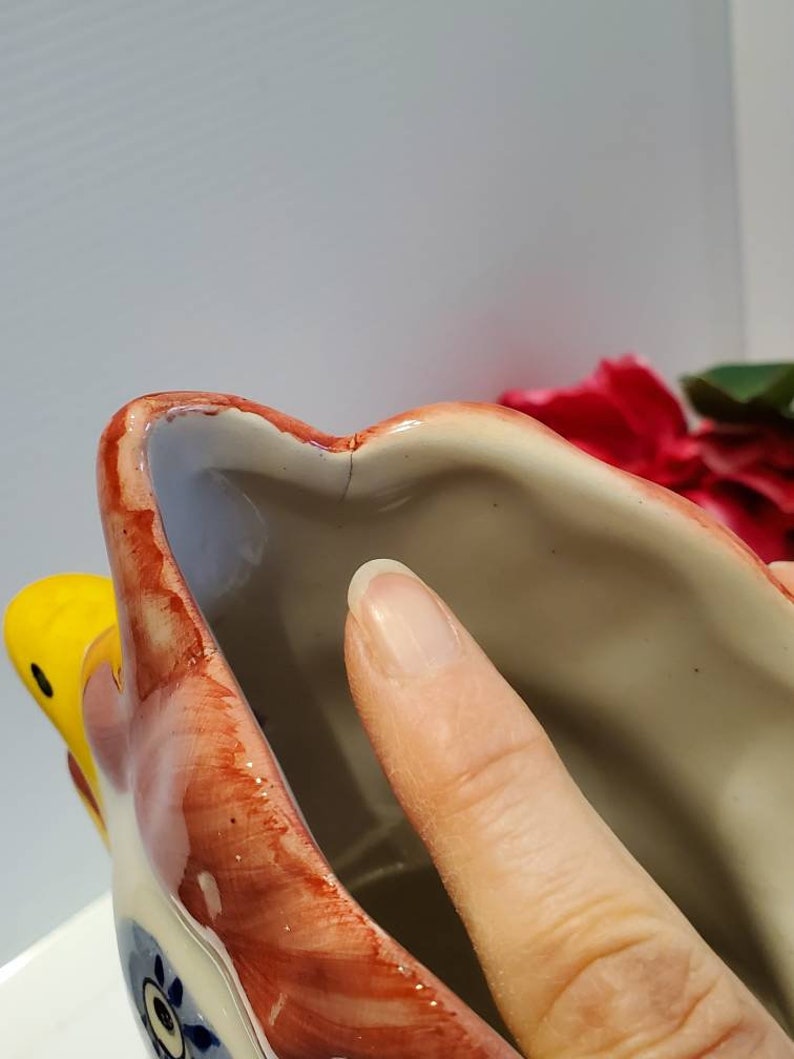 May include: Close-up of the inside of a ceramic rooster planter. The planter is painted in shades of red, yellow, and blue. The inside of the planter is white.
