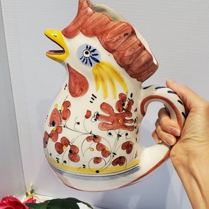 May include: A white ceramic pitcher shaped like a rooster with red, yellow, and blue painted details. The rooster has a large beak and a comb on its head. The pitcher has a handle and a floral design.