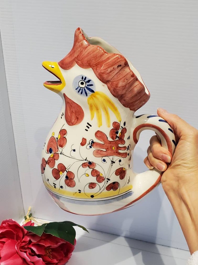 May include: A white ceramic pitcher shaped like a rooster with red, yellow, and blue painted details. The rooster has a large beak and a comb on its head. The pitcher has a handle and a floral design.