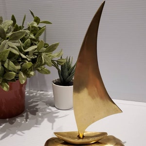 May include: A gold-colored metal sailboat sculpture with a raised base. The sailboat has a single sail and is a modern design.