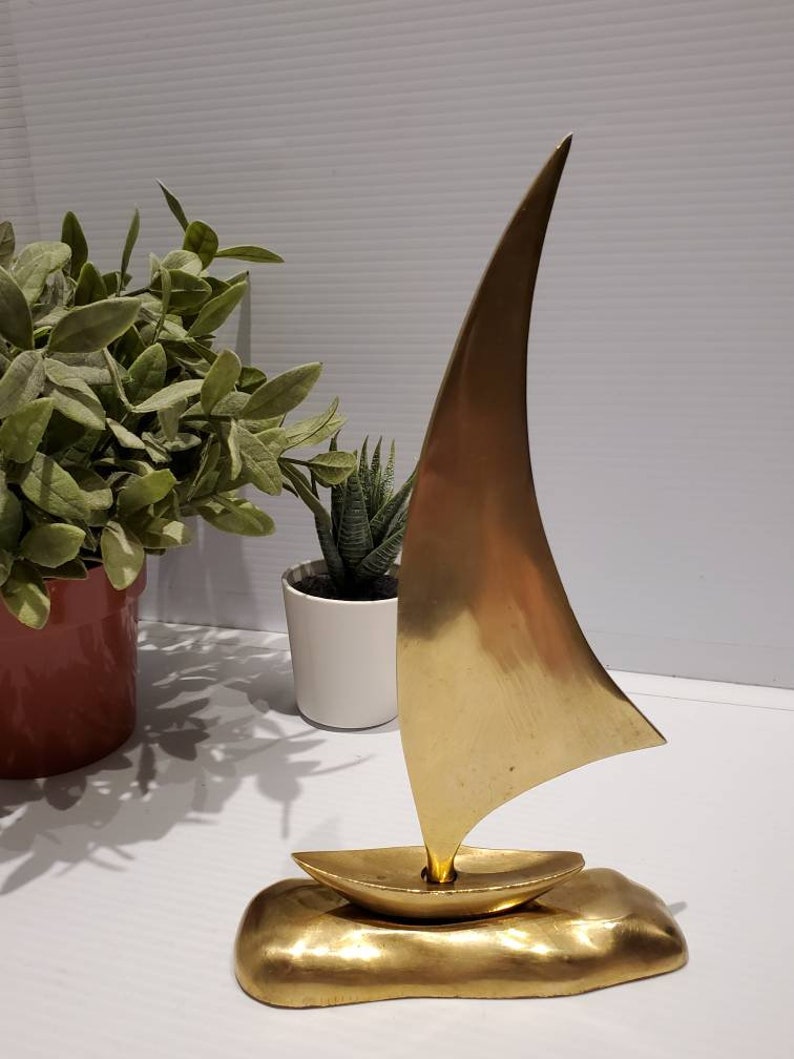 May include: A gold-colored metal sailboat sculpture with a raised base. The sailboat has a single sail and is a modern design.