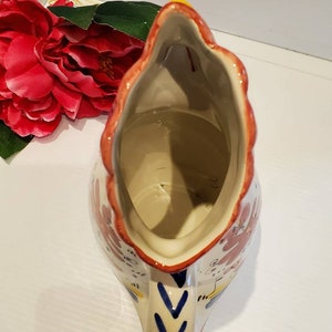 May include: A white ceramic pitcher with a rooster design. The pitcher has a red, yellow, and blue floral pattern. The pitcher is empty and the inside is visible.