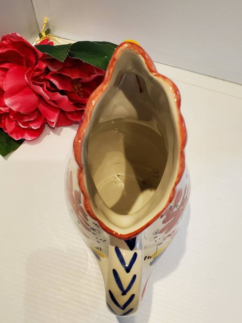 May include: A white ceramic pitcher with a rooster design. The pitcher has a red, yellow, and blue floral pattern. The pitcher is empty and the inside is visible.