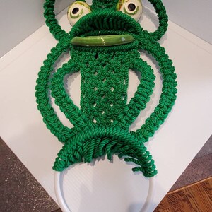 May include: Green crocheted frog towel ring with white ceramic eyes and a green ceramic mouth. The ring is attached to a white metal ring.