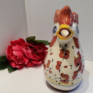 May include: A white ceramic pitcher shaped like a rooster with red, yellow, and blue accents. The pitcher is decorated with a floral pattern in shades of red and yellow.