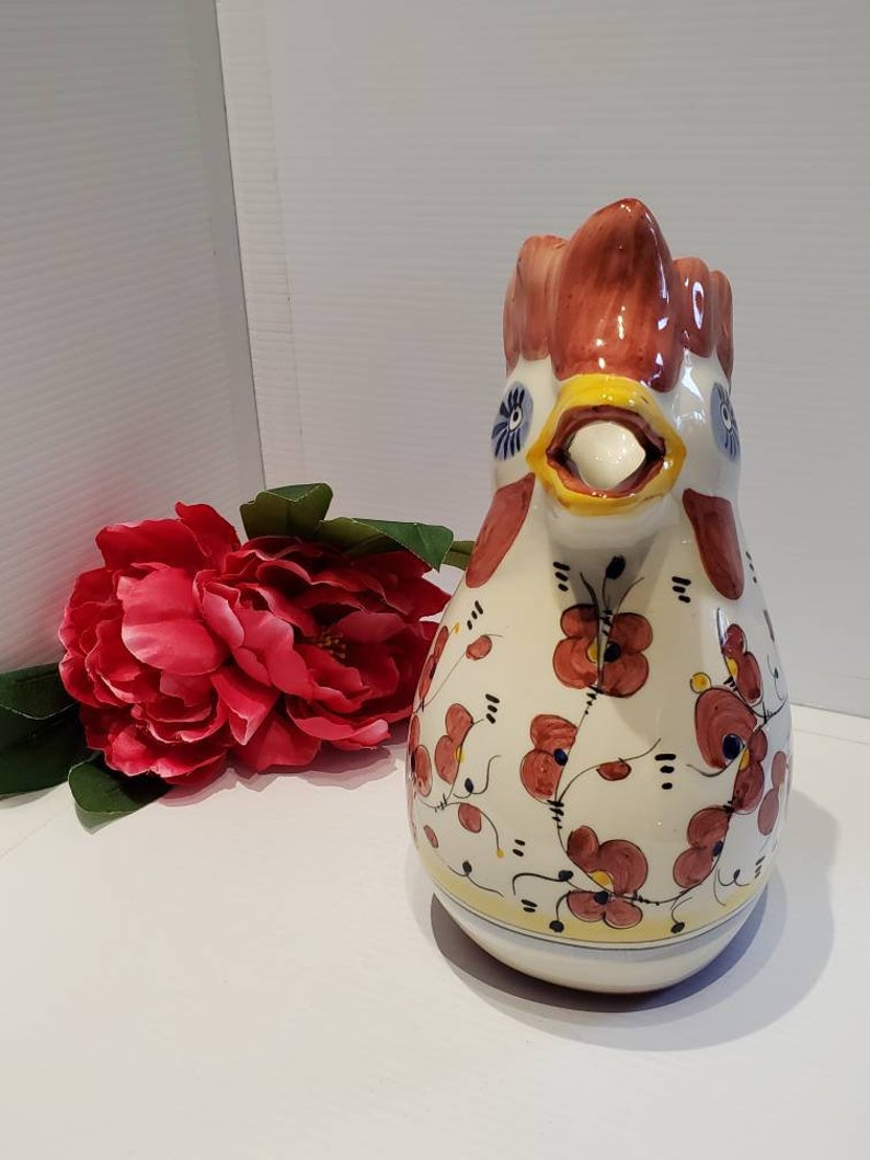 May include: A white ceramic pitcher shaped like a rooster with red, yellow, and blue accents. The pitcher is decorated with a floral pattern in shades of red and yellow.