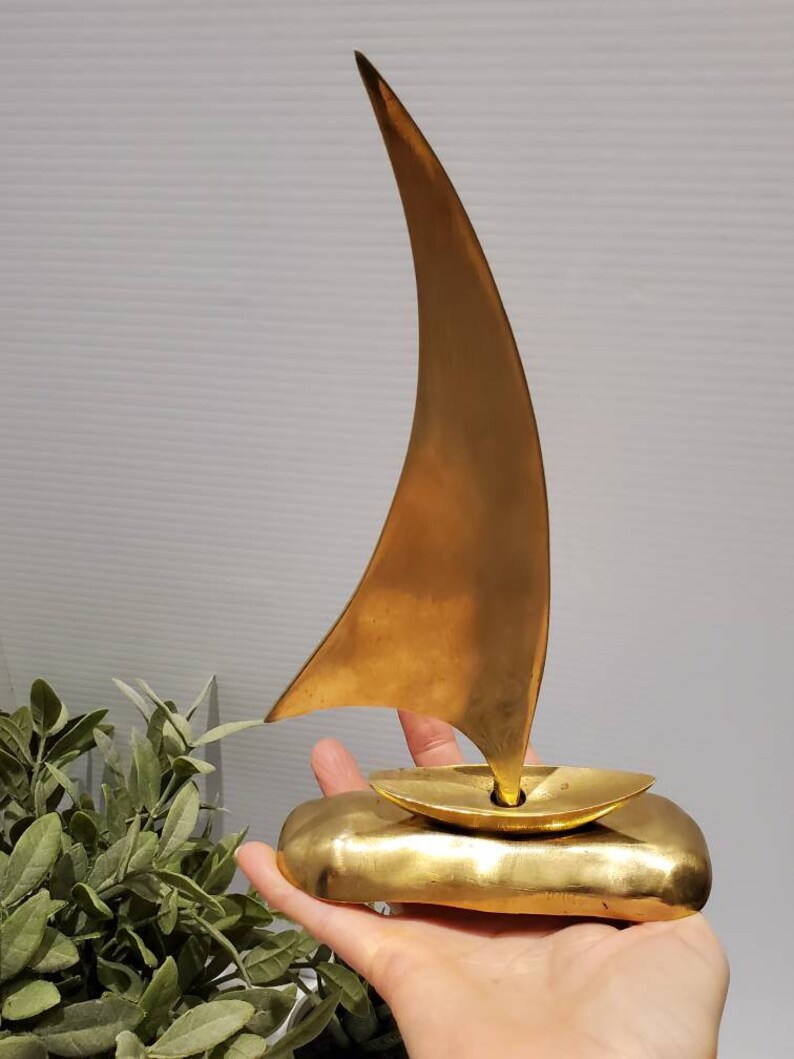 May include: A gold-colored brass sailboat sculpture with a single sail. The sailboat is sitting on a base that resembles a wave.