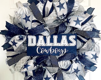May include: A decorative wreath featuring a Dallas Cowboys theme. The wreath is made with navy blue, silver, and white ribbons, with star accents. A navy blue sign in the center displays the words "DALLAS Cowboys".