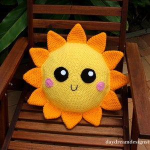 May include: A yellow crocheted sun pillow with a smiling face, black eyes, and pink cheeks. The pillow has orange crocheted rays.