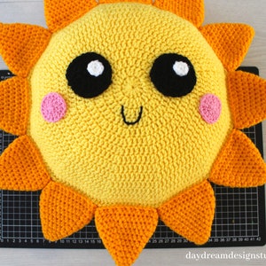 May include: A crocheted sun pillow with a yellow face, orange rays, and black eyes. The sun has pink cheeks and a smiling mouth.